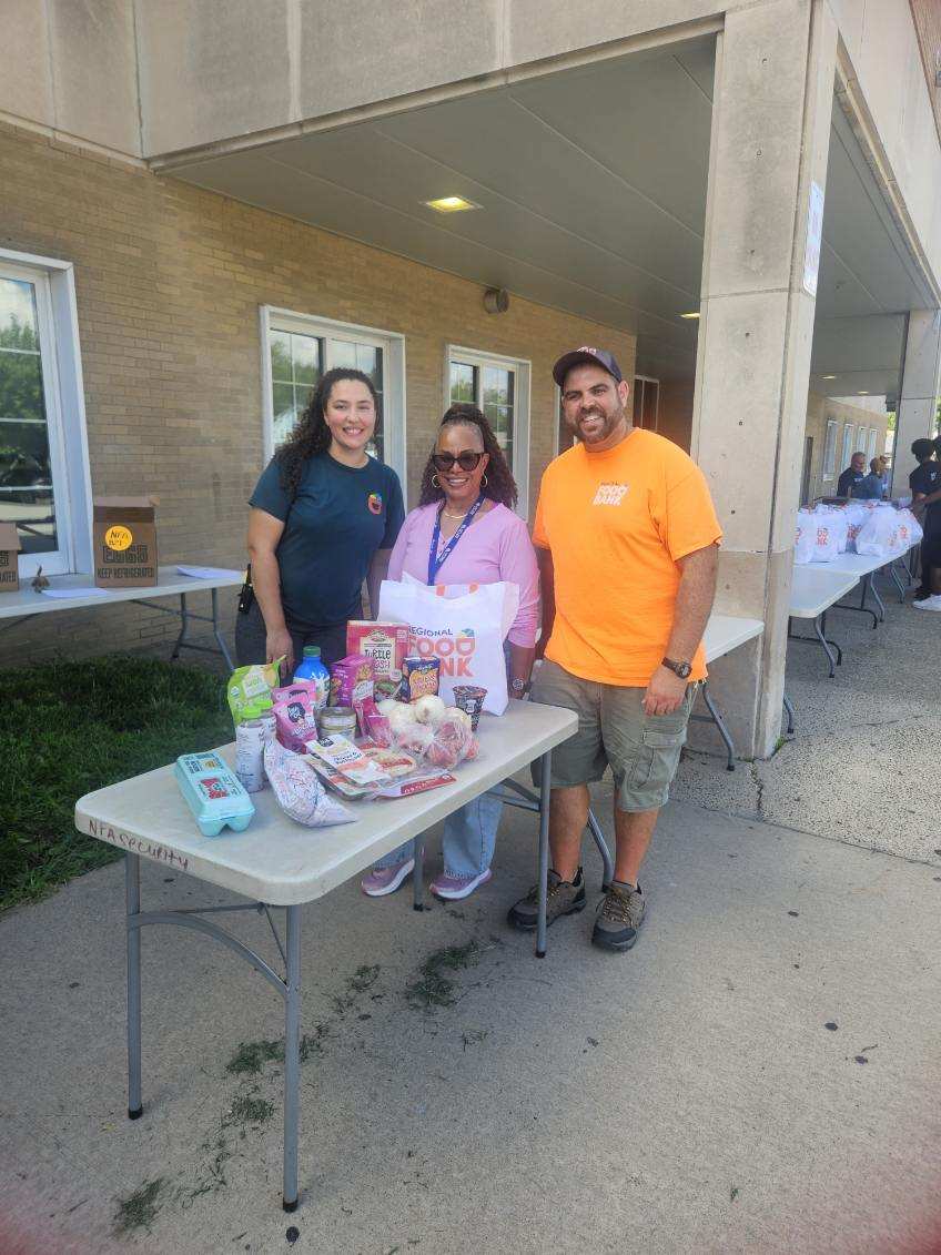 Three people stand behind a table of donated food items outside a building.