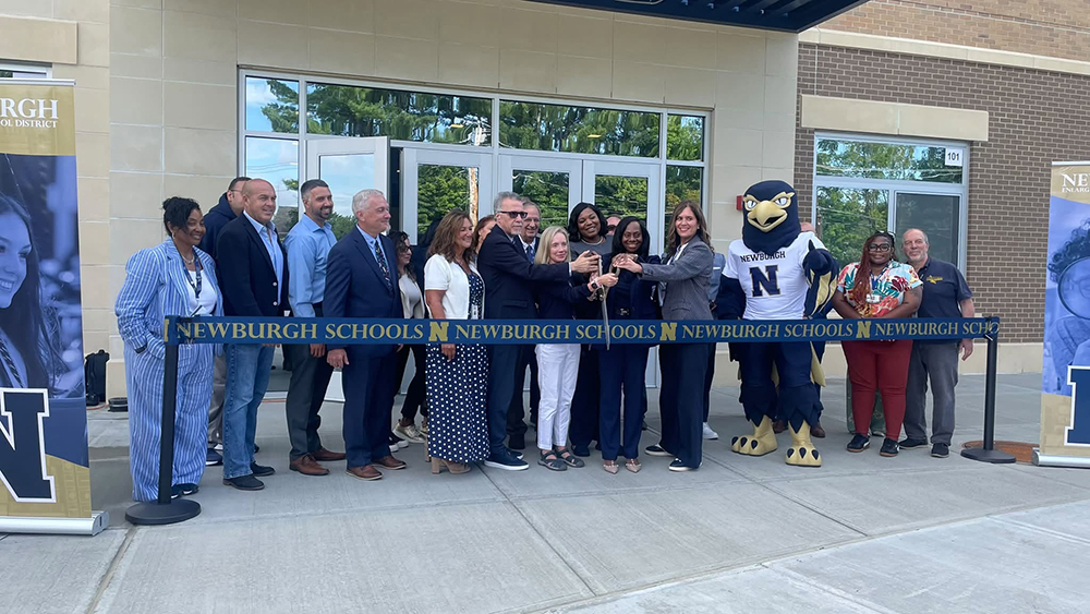 A group of people pose outside a new school building during a ribbon-cutting ceremony with the Newburgh school mascot.