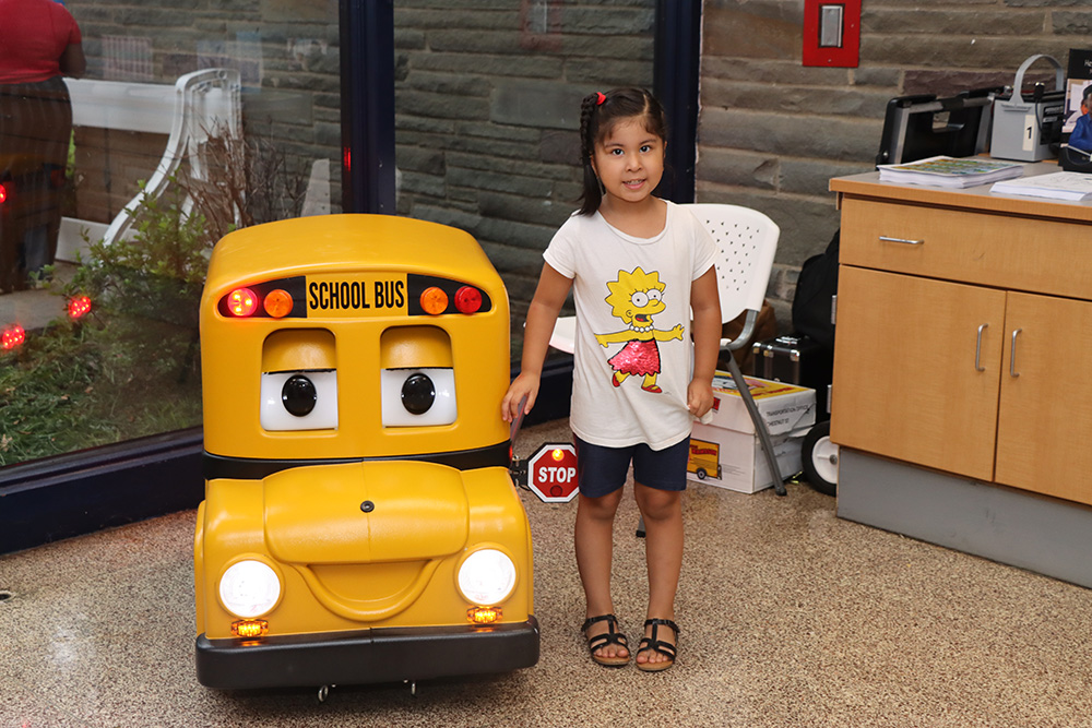 A young child stands beside a small yellow school bus display inside a school building.